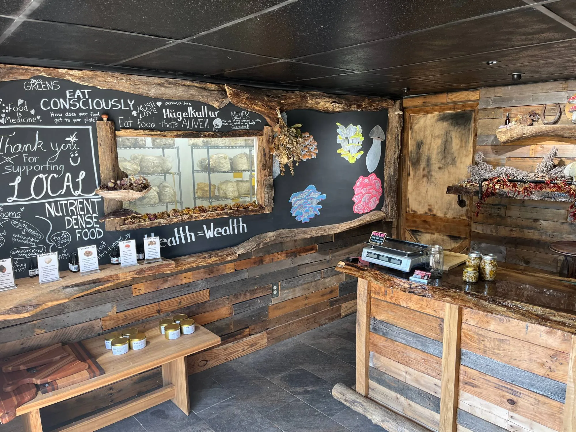 Inside the Mush Love shop: a chalkboard wall framed in live-edge wood reading “Thank you for supporting LOCAL”, a window into the grow room, tincture displays and a live-edge wooden counter